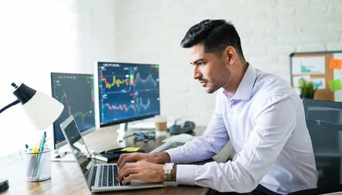 Right investment strategy - tycoonstory | tycoonstory media A man working at his desk analyzing stock charts on dual monitors while typing on a laptop in a modern office setting.