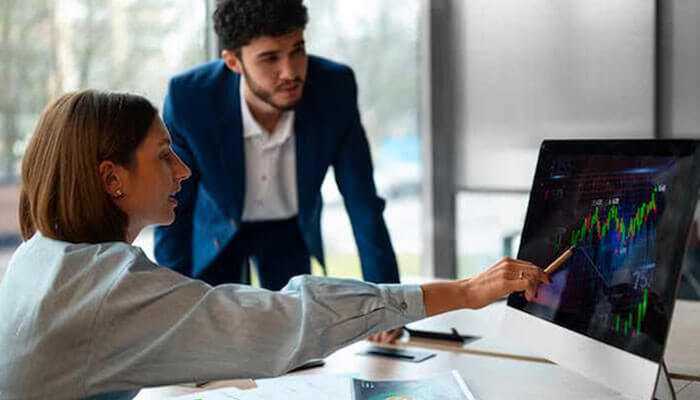 Two professionals analyze stock market charts together in an office setting, discussing smart investing trends and data. Two professionals analyze stock market charts together in an office setting, discussing smart investing trends and data.