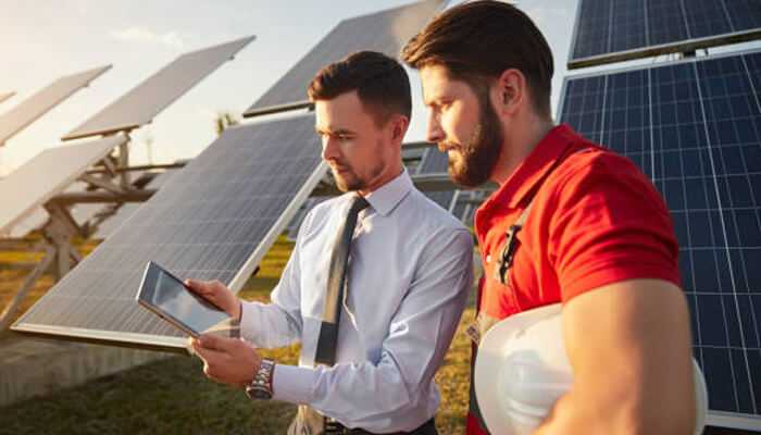 Understanding solar panel cost in delhi - tycoonstory | tycoonstory media Two professionals discussing solar panel installation at a solar farm, with one holding a tablet and the other wearing a safety helmet.