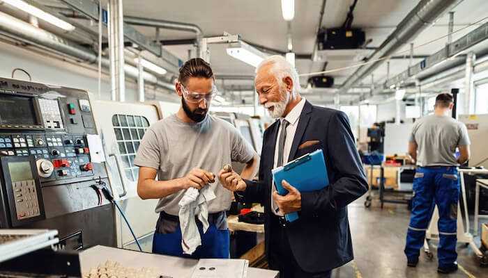 A company that handles everything for you | tycoonstory media An engineer discussing a machine part with a manager in a factory setting — representing the process to hire a mechanical engineer for manufacturing projects.