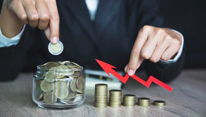 Achievable financial goals | tycoonstory media A person placing coins in a jar and pointing at a downward red arrow with stacked coins, symbolizing financial challenges and steps toward financial prosperity.