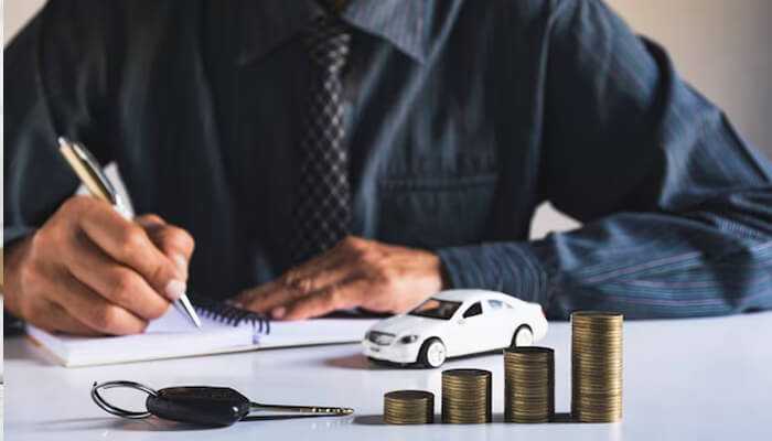 Average car loan interest rates for pre-owned | tycoonstory media Car loan interest rate concept – a businessman writing on a notepad with a car key, stacked coins, and a miniature car model on a table, representing auto loan financing and interest rates.