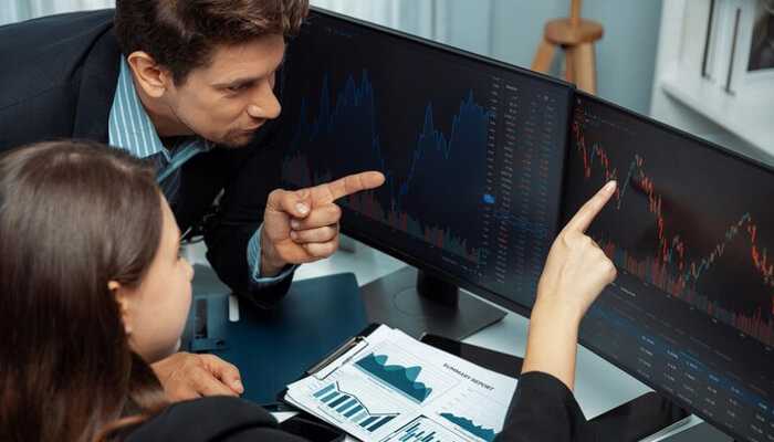 Model development and backtesting | tycoonstory media Quant researchers analyzing financial market charts on dual monitors, discussing backtesting results and model performance metrics.