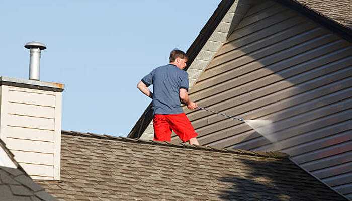 Pressure wash the siding | tycoonstory media A man cleaning shingles on a roof as part of exterior home improvement work.