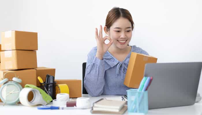 Product | tycoonstory media Young female entrepreneur managing her small e-commerce business, smiling while packing products and engaging with customers online, showcasing the growing impact of digital platforms on small businesses.
