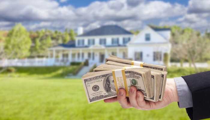 Speed and accessibility | tycoonstory media A hand holding stacks of hundred-dollar bills in front of a large house, symbolizing fast access to funds through a hard money loan for real estate investment.