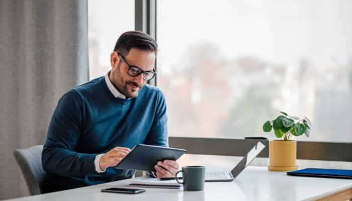 The benefits of taking a break | tycoonstory media A man sitting at a desk using a tablet, with a relaxed demeanor and a calm work environment, reflecting a stress-free approach to managing tasks.