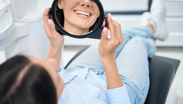 Tooth replacement | tycoonstory media Woman smiling while looking at her reflection after dental treatment, representing the satisfaction achieved with dental implants vs bridges solutions.
