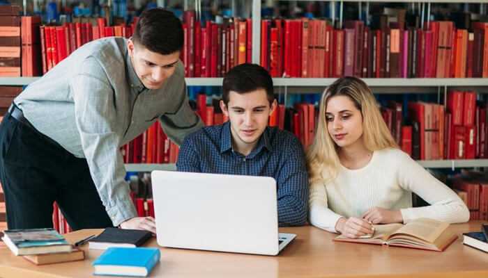 Top skills required for a business management career | tycoonstory media Three students studying together in a library, researching career opportunities in business management in india using a laptop and books.