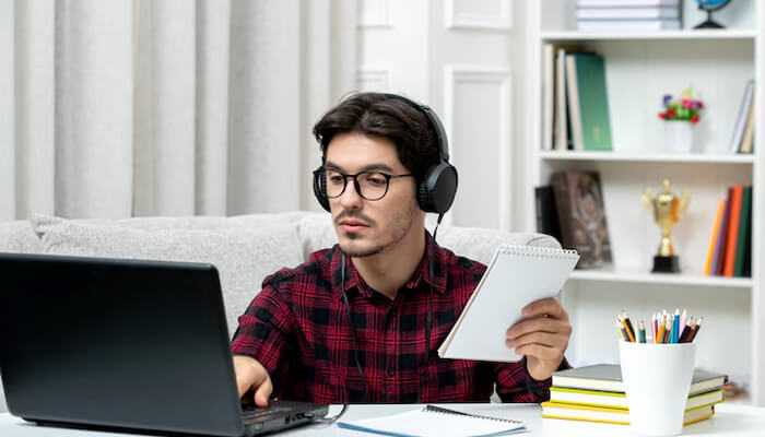 | tycoonstory media Young man wearing headphones and glasses, working on a laptop and holding a notebook, representing employer of record for global hiring and remote workforce management.