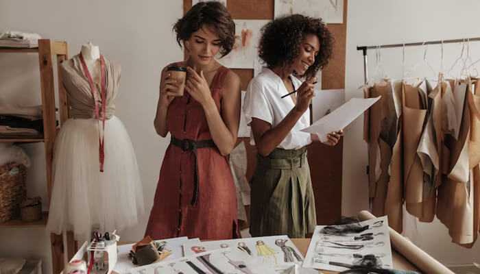 Building unique outfits with what you have | tycoonstory media Two women brainstorming fashion sketches in a design studio with mannequins and fabrics, exploring creative women’s outfits ideas using existing wardrobe pieces.