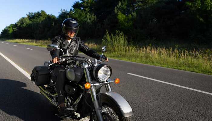 Helmets weather and what to expect 1 | tycoonstory media Motorcyclist wearing a helmet and leather jacket riding a motorcycle on a clear road surrounded by green trees, representing the experience of holding a wyoming motorcycle license.