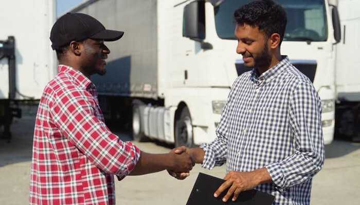 How staffing agencies are revolutionizing driver recruitment | tycoonstory media Two men shaking hands near trucks, symbolizing collaboration and recruitment innovations in trucking industry trends.