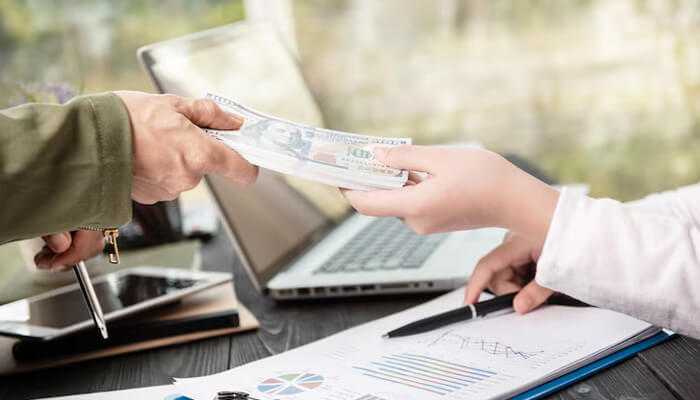 Increasing access for underrepresented groups | tycoonstory media Close-up of hands exchanging cash over financial charts and a laptop, representing real-world access to pedirayudas government financial support initiatives.