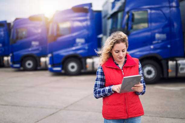 Istockphoto-1189954840-612x612 | tycoonstory media A woman in a red vest using a digital tablet in front of a row of blue trucks, representing modern technology adoption in trucking industry trends.