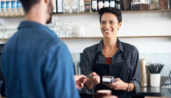 Payment flexibility of payment choice | tycoonstory media A smiling barista handing a payment terminal to a customer, showcasing flexible payment methods and secure payments for small businesses.
