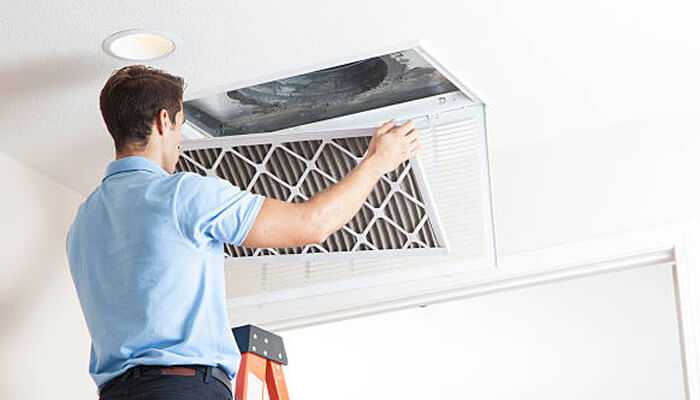 Seasonal considerations for filter changes | tycoonstory media Man on ladder conducting furnace filter replacement by installing a new air filter in a ceiling hvac vent to improve indoor air quality.