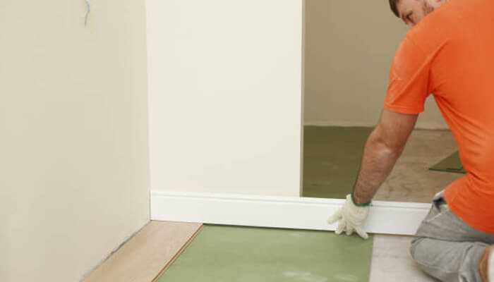 Strong structure and easy setup | tycoonstory media Installer fitting a white skirting board along the base of a wall over green floor underlayment.