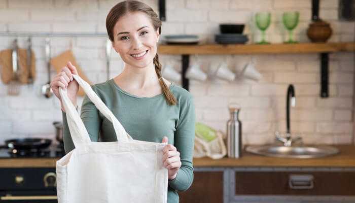 Sustainability | tycoonstory media A woman smiling while holding a custom tote bag in her kitchen, showing a practical and eco-friendly accessory for everyday use.