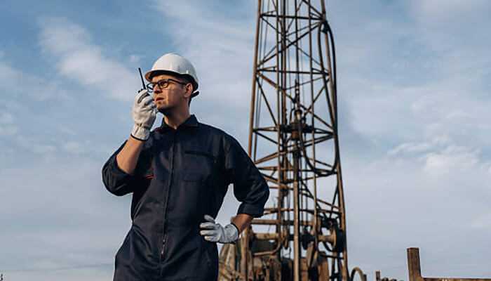 The role of frequency in walkie talkie range | tycoonstory media Technician in safety helmet using poclink walkie talkie near an industrial drilling rig under blue sky.