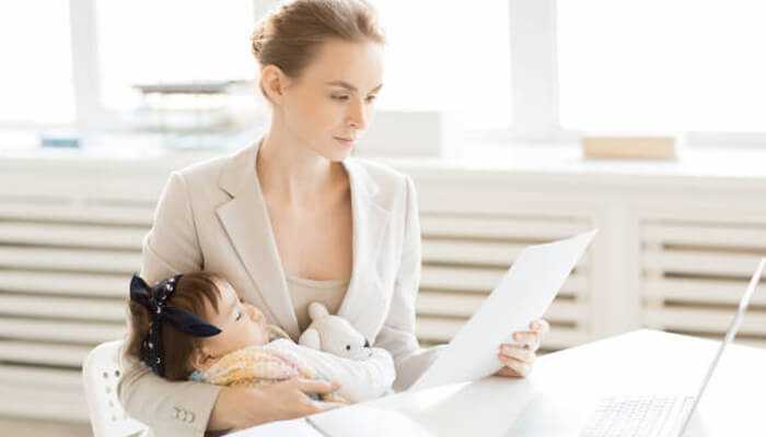 Add on-site or subsidized childcare options 1 | tycoonstory media Working mother holding her baby while reading documents at her desk, showing family friendly benefits at work.