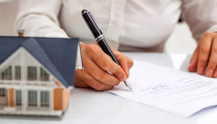 Carefully evaluate loan options | tycoonstory media Close-up of a person signing home purchase documents next to a miniature house, symbolizing the process of buying a house.