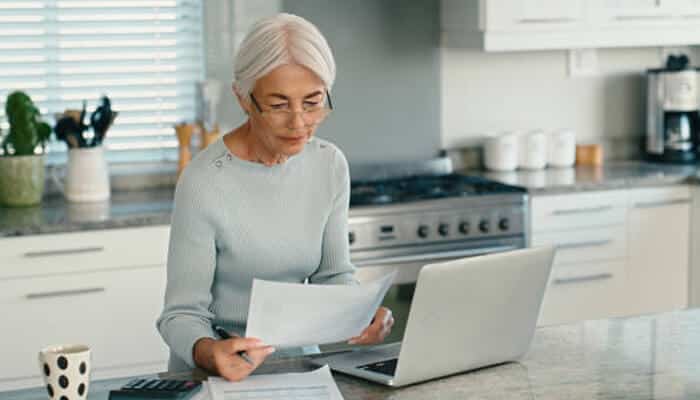 Factors to consider when reviewing it | tycoonstory media Senior woman reading life insurance policy documents at home while using a laptop to manage financial planning.