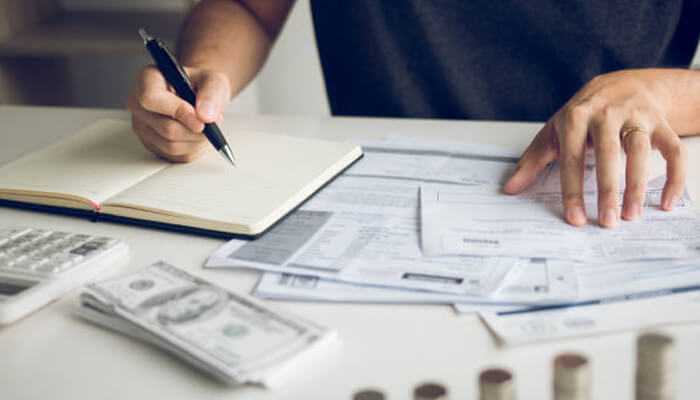 Failing to record income and expenses | tycoonstory media A person working on finances and tax mistakes with a notebook, calculator, bills, and stacks of money on the desk.