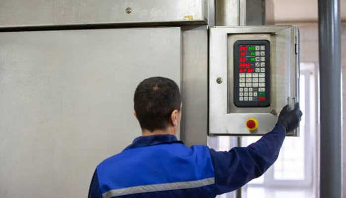 How can we improve the accuracy of measurements | tycoonstory media Worker adjusting a control system with load cells in a factory setting, interacting with a machine panel for operations monitoring.