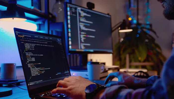 Reduces dependency on outsourced technical talent | tycoonstory media A close-up of a person coding on a laptop in a modern workspace, with dual screens displaying code, illustrating the role of technical education in software development and programming.