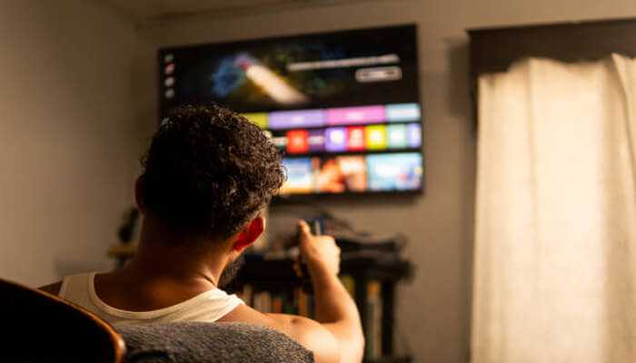The technology behind nigma | tycoonstory media A person sitting on a chair, holding a remote control and watching nigma streamz iptv on a smart tv, exploring streaming content.