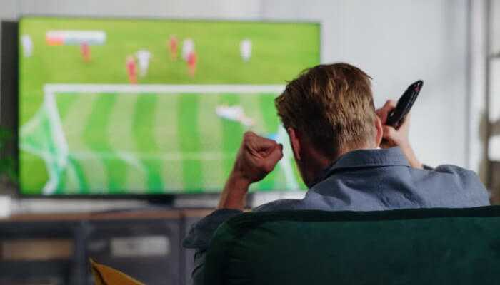 Underrated sites worth your attention | tycoonstory media A man watching a soccer match on tv at home, excitedly raising his fist while holding a remote, illustrating how to watch free live sports.