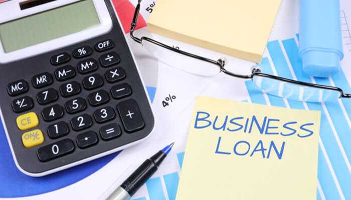 What are the terms like | tycoonstory media Calculator, eyeglasses, and a yellow sticky note reading “business loan” placed on financial charts, representing commercial loans and financing tools