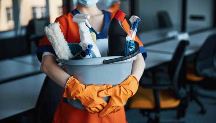 Check for efficiency and versatility | tycoonstory media A person wearing protective gloves, holding a bucket filled with commercial cleaning products like sprays, brushes, and cleaning solutions in an office setting.