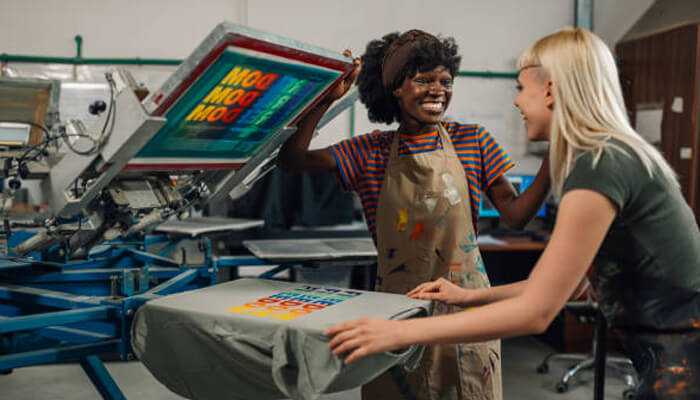Custom merchandising Two women working together in a custom merchandising workshop, with one holding a screen-printed t-shirt featuring a colorful design. The process showcases the creative side of custom merchandise production.