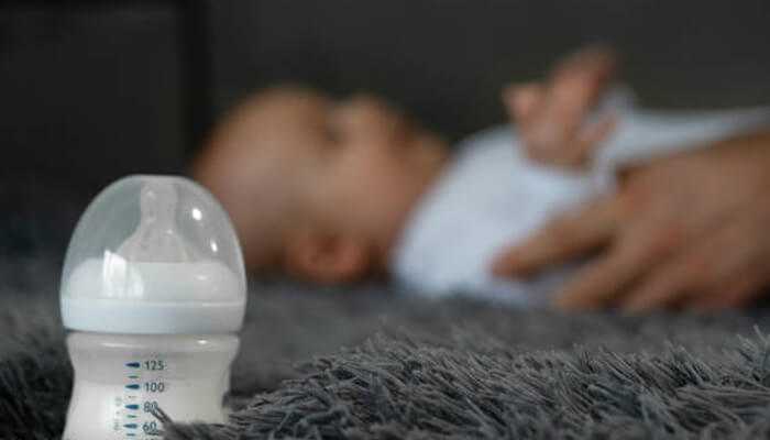 Could it be a feeding schedule issue | tycoonstory media A close-up of a baby bottle filled with milk placed on a soft carpet, with a baby lying in the background, highlighting the challenge of a baby refusing the bottle.