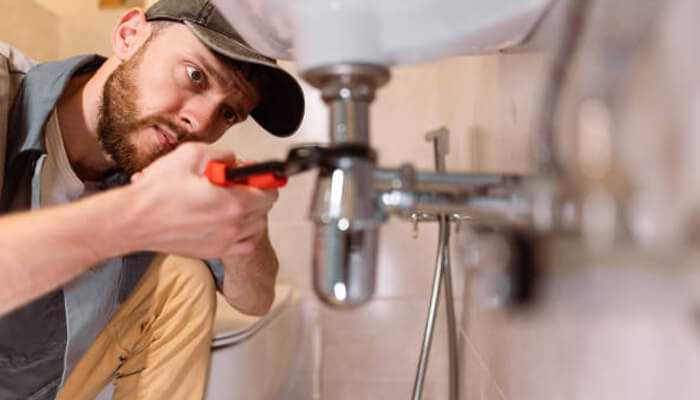 Plumber | tycoonstory media A skilled trades professional using a wrench to repair plumbing under a bathroom sink.