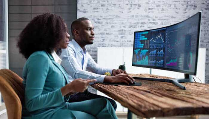 Real-time data Two professionals analyzing real-time data on a large curved monitor in a modern office setting. One is using a smartphone while the other is focused on the data analysis displayed on the screen, showcasing financial and market trends.