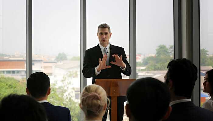 The impact of motivational speakers in corporate settings 1 | tycoonstory media Engaging motivational speaker in a business suit speaking energetically to an audience in a bright, nature-filled indoor setting.