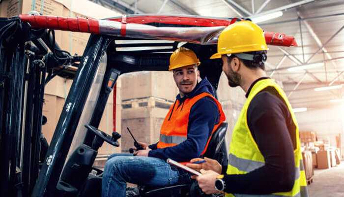 The power of third-party safety consulting | tycoonstory media Two warehouse workers discussing procedures near a forklift, emphasizing communication and awareness of safety risks for warehouses.