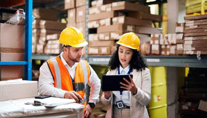 Types of warehousing services 1 | tycoonstory media Two warehouse professionals in safety gear reviewing data on a tablet, highlighting the teamwork and efficiency of warehousing services.
