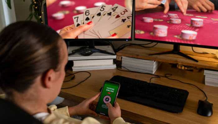 What to know before playing poker online for real cash | tycoonstory media Woman playing poker online using her smartphone while watching live cards and chips on dual monitors in a home gaming setup.