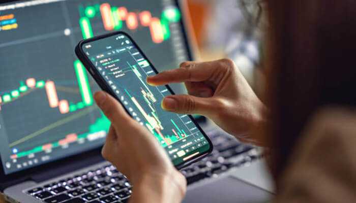 Broad market access | tycoonstory media Person using the zengtp mobile trading app on a smartphone with stock charts displayed on a laptop in the background.