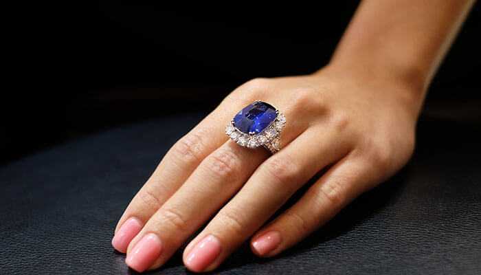 Colour craftsmanship and emotional resonance - tycoonstory | tycoonstory media Elegant sapphire engagement ring with a vibrant blue gemstone and diamond halo, worn on a woman’s hand with manicured nails.