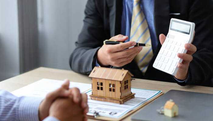 Legal fees and closing costs 1 | tycoonstory media Mortgage consultation scene showing a broker using a calculator while discussing home loan details with a client beside a small wooden house model.