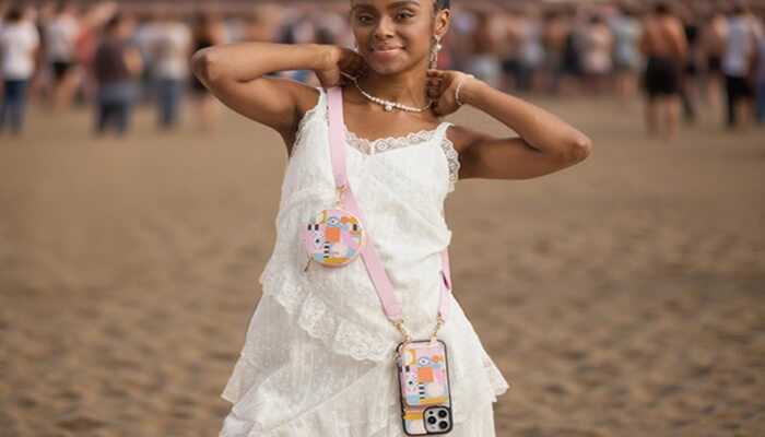 Crossbody wallet phone case
A smiling young woman, dressed in a layered white lace dress, stands on a sandy beach or open field with a crowd of people blurred in the background. She is wearing a pearl choker necklace and holding a pose with her hands near her head.