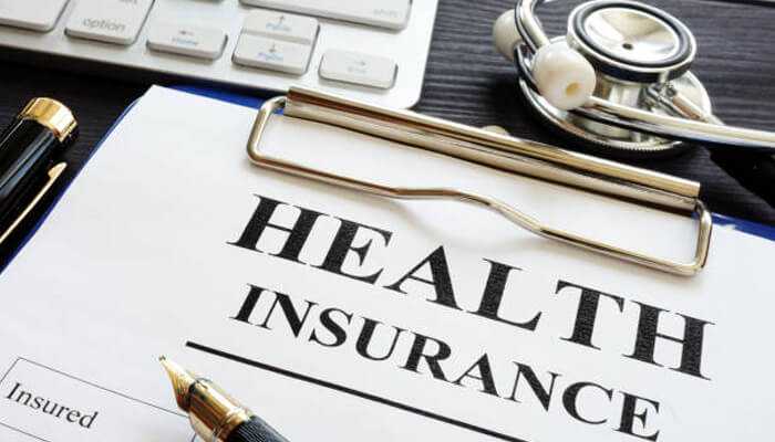 Health insurance A document on a clipboard with the bold words " health insurance " printed across the center. A black pen with a gold nib and a stethoscope rest on the document, and a computer keyboard is visible in the background.