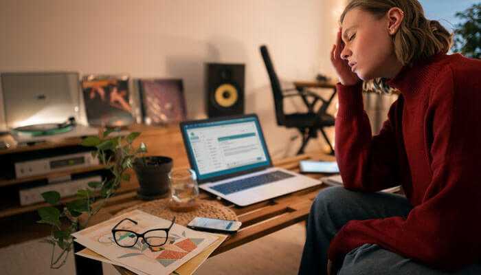 How remote work influences mental health policies - tycoonstory | tycoonstory media A woman working from home looking stressed in front of her laptop, highlighting the importance of updating employee health policies to support mental well-being.