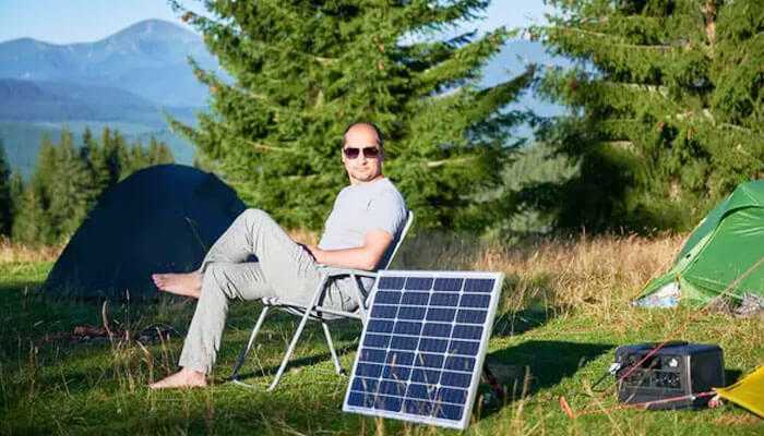 Choosing the right solar generator and portable power station - tycoonstory | tycoonstory media A man relaxing in a camping chair beside a tent with portable solar power generators harnessing sunlight in the mountains.