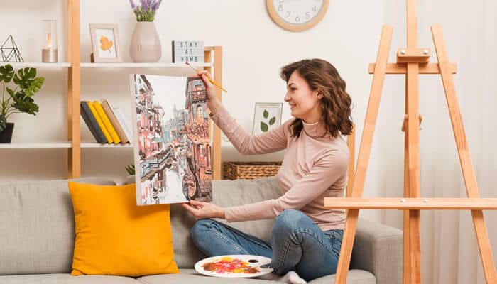 Artistic wall art and model design - tycoonstory | tycoonstory media A smiling woman sitting on a couch at home, holding up a canvas she is painting with a brush, with an easel and an artist's palette in the foreground.